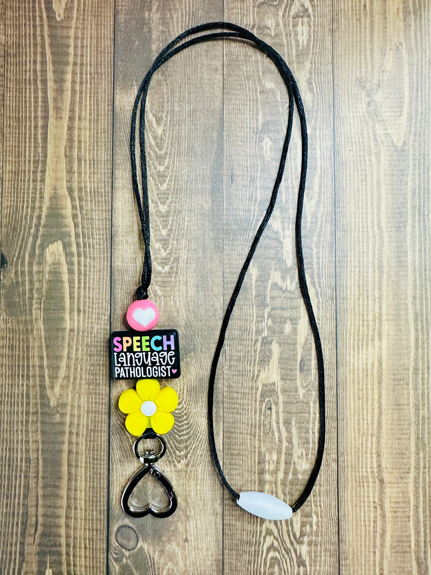 Full-length speech language pathologist lanyard laid flat on wooden table showing SPEECH focal bead, pink heart bead, and yellow flower bead.