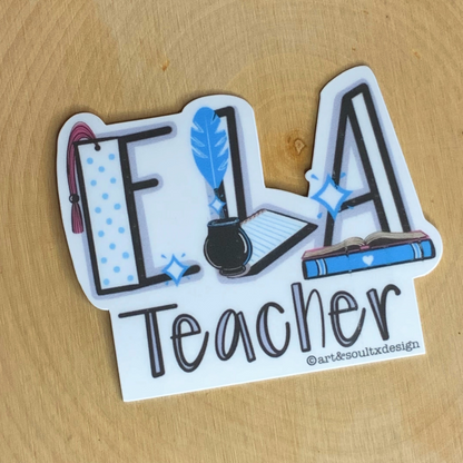 The ELA Teacher sticker has a bookmark, quill and ink, and open book and is laying on a wooden teacher desk.